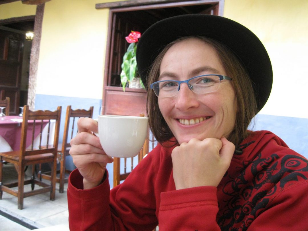 In the imagie is me - a white woman with long brownish hair and blue glasses. Im sitting at a table, holding a coffee cup and smiling in the camera. I have a black hat on my head.